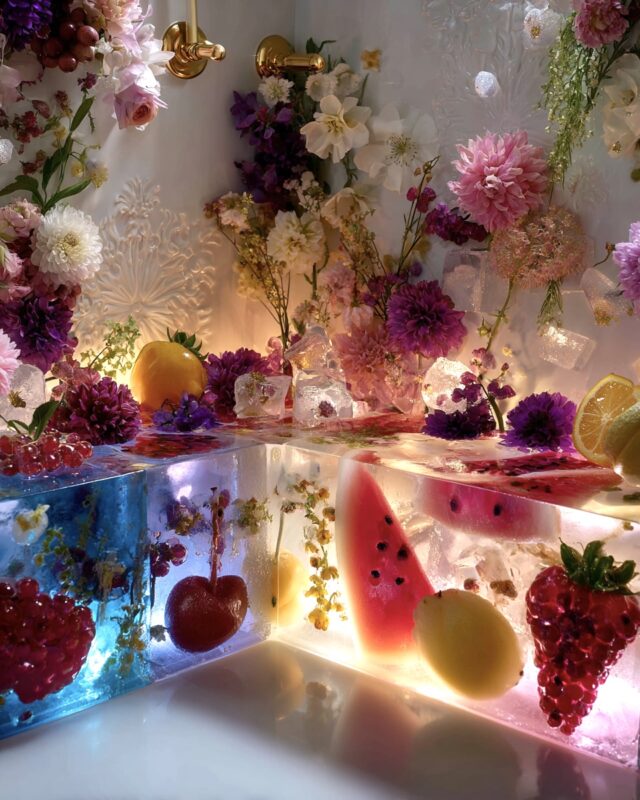 Corner shower bench made of glowing translucent resin blocks embedded with fruit slices and flowers, surrounded by gold fixtures and blooming florals.